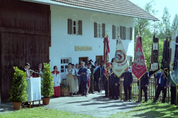Bei der Museunseröffnung steht Heinrich Maul am Rednerpult vor dem Baschtlehaus, daneben steht Pfarrer Hiebsch, ein Ministrant und Fahnenabordnungen der örtlichen Vereine